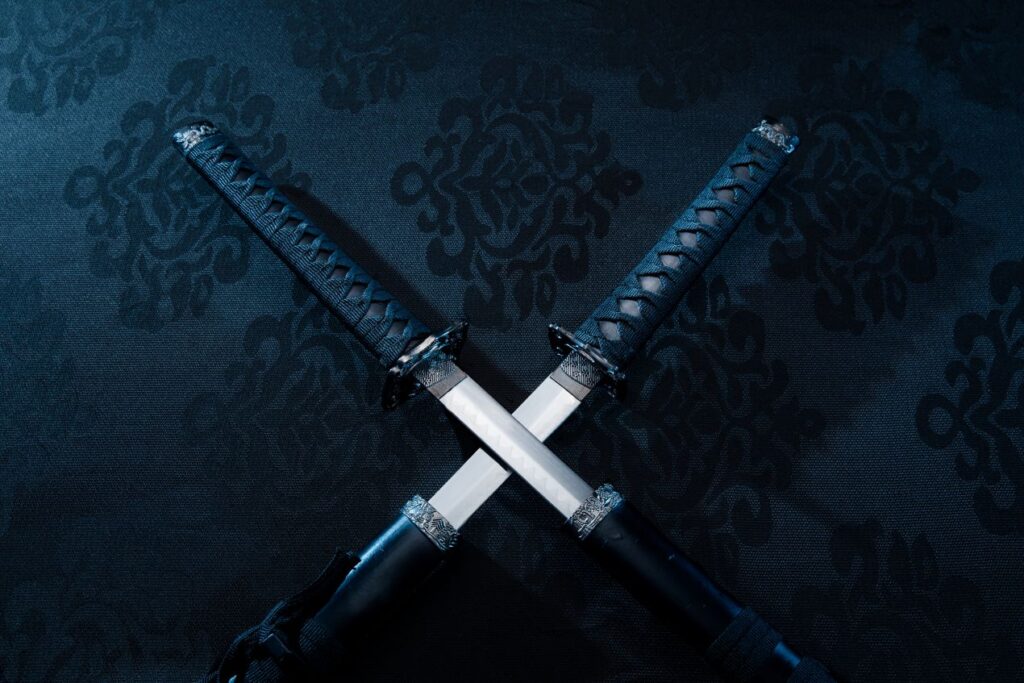 Zwei gekreuzte Katana mit verzierten Griffen auf dunklem Hintergrund, symbolisieren Tradition, Kampfkunst und japanische Schmiedekunst.